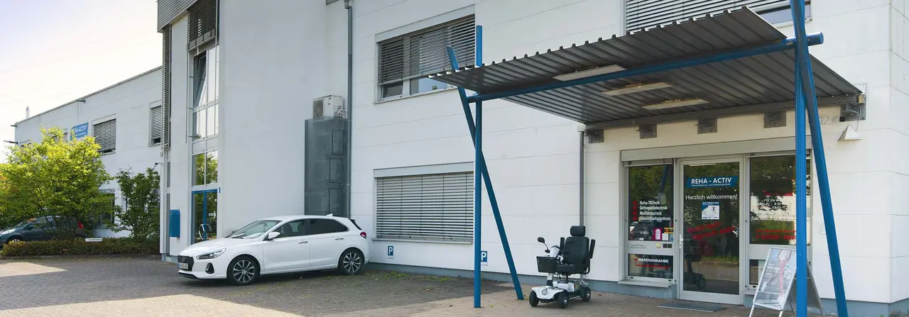 Außenansicht des Sanitätshauses Reha-Activ in Röstrath: weißes Gewerbegebäude mit verglastem Eingangsbereich unter einem Vordach auf blauen Stahlstützen, davor ein Elektromobil auf dem Gehweg und ein Parkplatz mit einem weißen PKW.