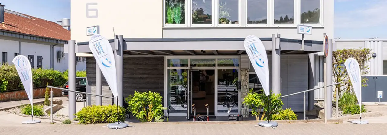 Außenansicht des Sanitätshaus Lammert & Scherer in Wörstadt: modernes Ladengebäude mit überdachtem Eingang, vier weißen Beachflags mit Firmenlogo, Rollator und Fahrrad vor der Glastür.