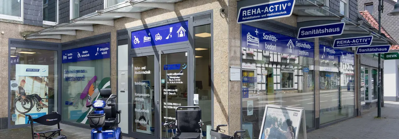 Außenansicht des Sanitätshauses Reha-Activ in Overath mit blau-grauer Fassade, vor dem Eingang ein ausgestellter Elektroroller und ein Rollstuhl.