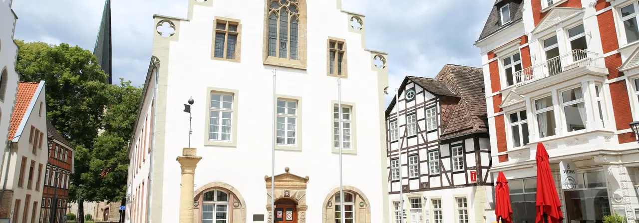 Ein historischer europäischer Stadtplatz mit weißen Fachwerkhäusern, roten Sonnenschirmen und einem Kirchturm im Hintergrund.