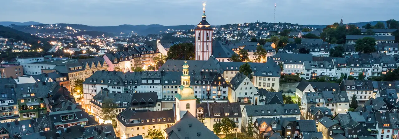 Luftaufnahme einer europäischen Stadt in der Abenddämmerung mit einem markanten Kirchturm, beleuchteten Straßen und den umliegenden Hügeln im Hintergrund.