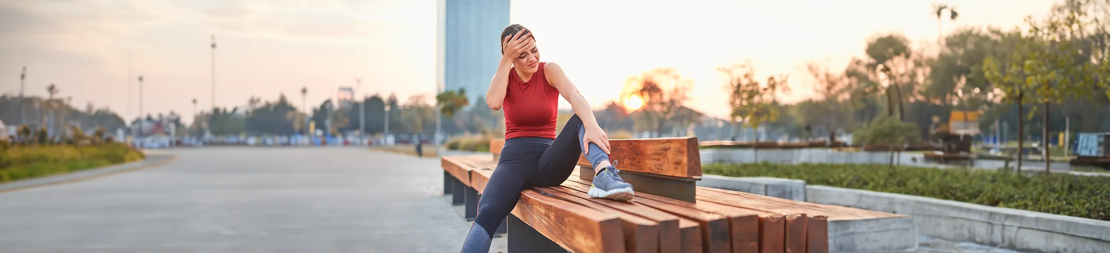 Bänderdehnung, Frau draußen auf Bank fasst sich an Fußgelenk, Schmerzen