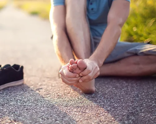 Mann mit Hohlfuß sitzt auf Boden draußen, hält sich den Fuß, Schuh steht daneben