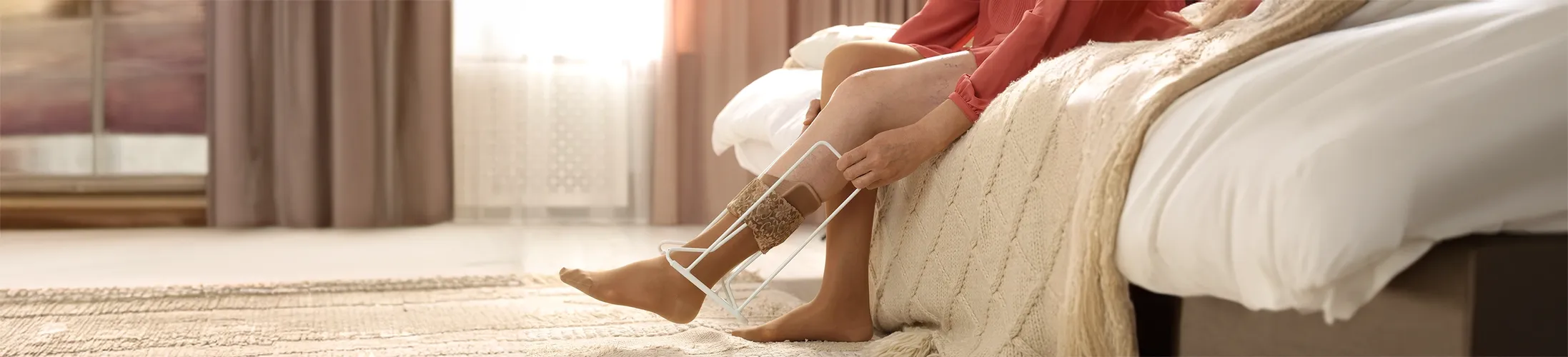 Kompressionsstrümpfe richtig anziehen, Frau sitzt auf Bett und benutzt eine Anziehhilfe