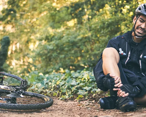 Füße schlafen beim Fahrradfahren ein, Mann sitzt neben Fahrrad und hält sich mit beiden Händen den Fuß,  Kribbeln in den Füßen, übermäßige Belastung führt zu Nerven- und Gefäßeinengungen