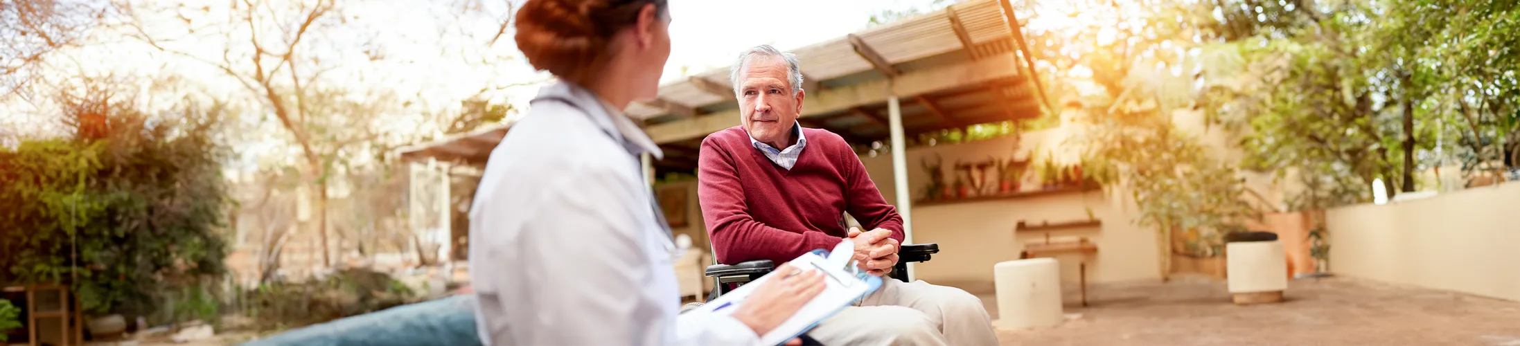 Mann sitzt im Rollstuhl, spricht mit Ärztin zu Kosten Rollstuhl, Bäume im Hintergrund