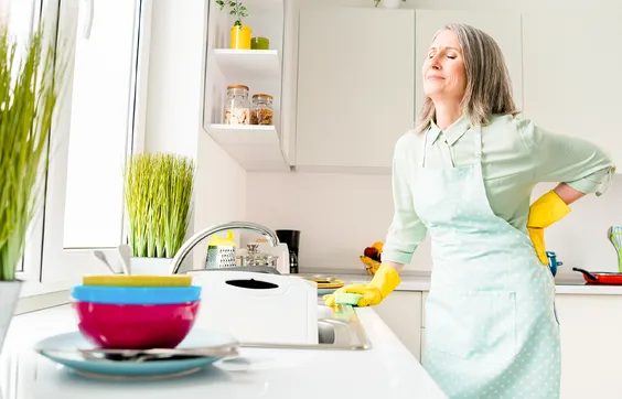 Frau hält sich beim Putzen den Rücken wegen Ischiasschmerzen, Küche mit Geschirr