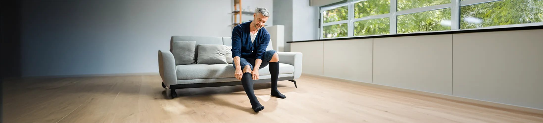 Kompression, Mann auf Sofa zieht sich Kompressionskniestrümpfe an, Vorbeugen gegen schwere Beine