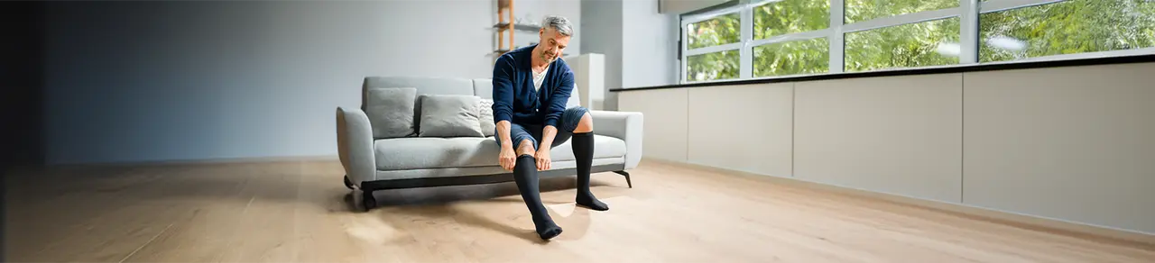Kompression, Mann auf Sofa zieht sich Kompressionskniestrümpfe an, Vorbeugen gegen schwere Beine