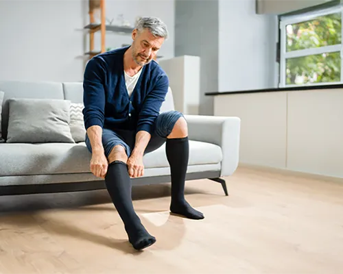Kompression, Mann auf Sofa zieht sich Kompressionskniestrümpfe an, Vorbeugen gegen schwere Beine