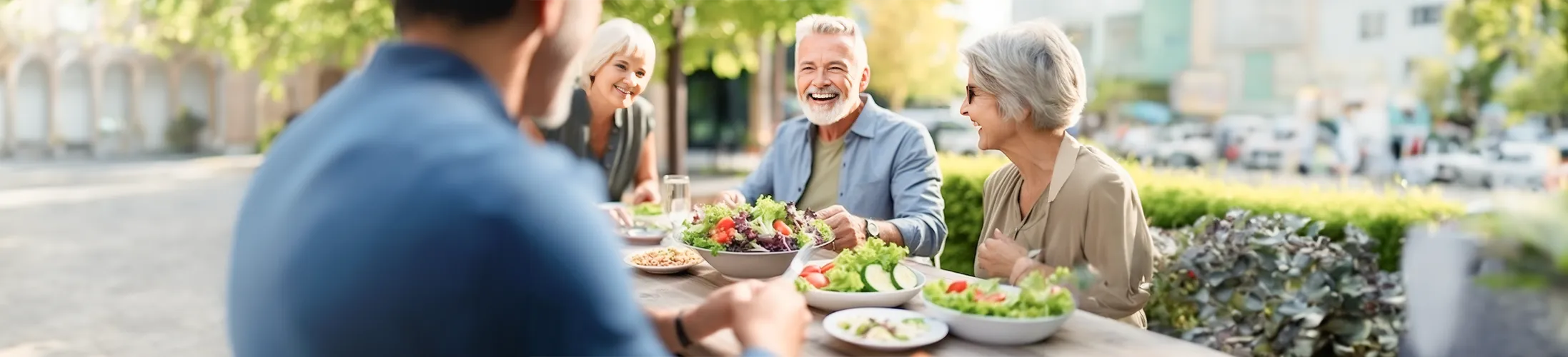 vier Personen mit Krampfadern sitzen an Tisch, essen Salate, Bäume im Hintergrund
