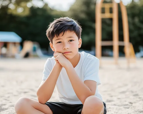 Junge mit Schürfwunde an Hand, sitzt auf dem Boden auf Spielplatz, schaut traurig