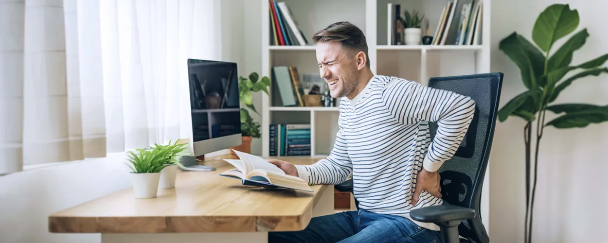 Mann sitzt auf einem Stuhl an einem Tisch, hält sich den Rücken mit schmerzverzehrtem Gesicht, hält ein Buch in der andere Hand, Pflanze im Hintergrund