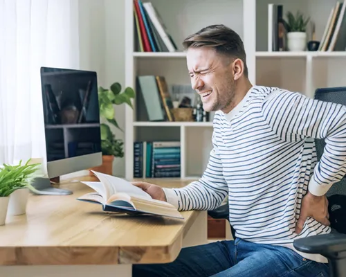 Mann sitzt auf einem Stuhl an einem Tisch, hält sich den Rücken mit schmerzverzehrtem Gesicht, hält ein Buch in der andere Hand, Pflanze im Hintergrund