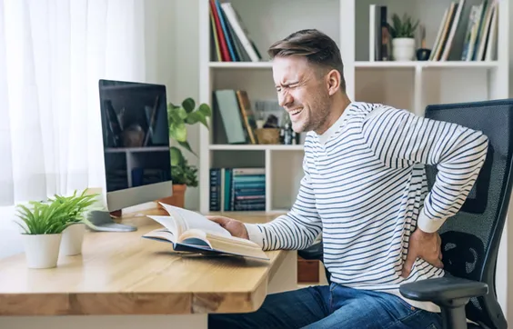 Mann sitzt auf einem Stuhl an einem Tisch, hält sich den Rücken mit schmerzverzehrtem Gesicht, hält ein Buch in der andere Hand, Pflanze im Hintergrund