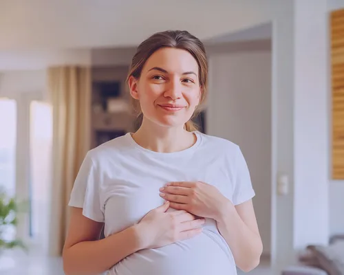 Schwangere Frau hält sich die Brust wegen Brustschmerzen, trägt weißes T-Shirt, Wohnung mit Sofia und Wandregal im Hintergrund