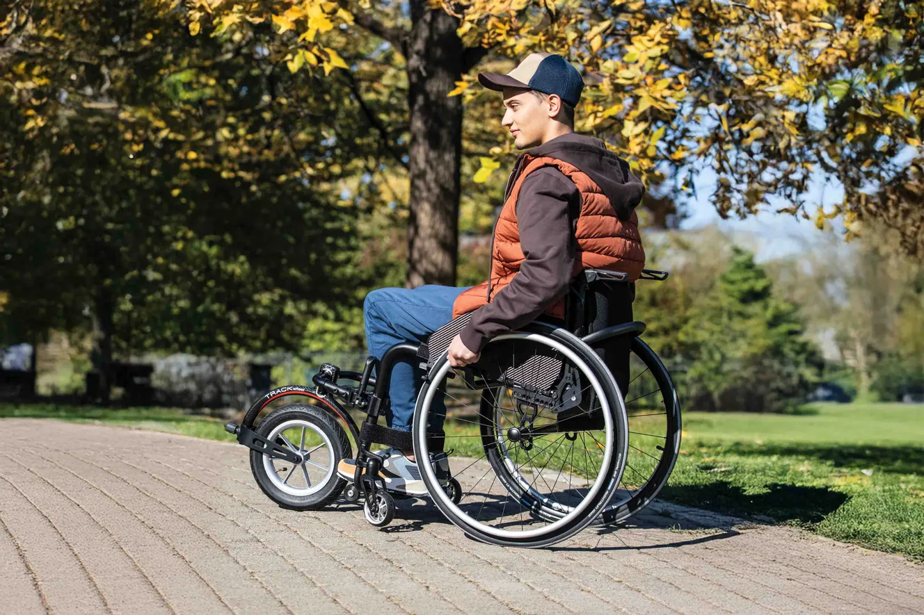 Mann sitzt im Rollstuhl mit Zuggerät von Rehasense, Track-Wheel, Vorderrad, steht in Park mit Bäumen, trägt rote Weste
