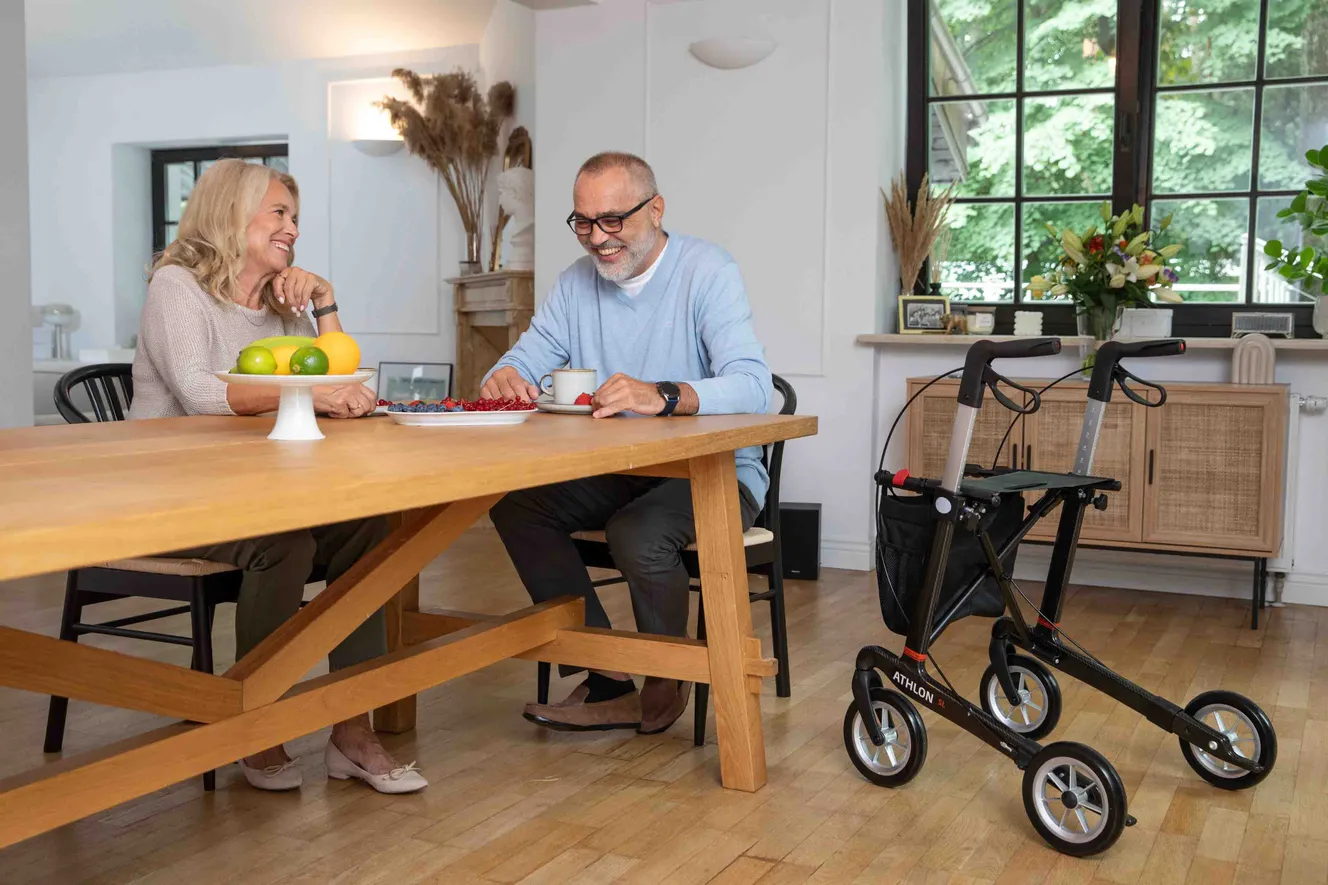 Pärchen sitzt in Wohnung an Tisch, Athlon-Rollator von Rehasense steht daneben, unterhalten sich, lachen