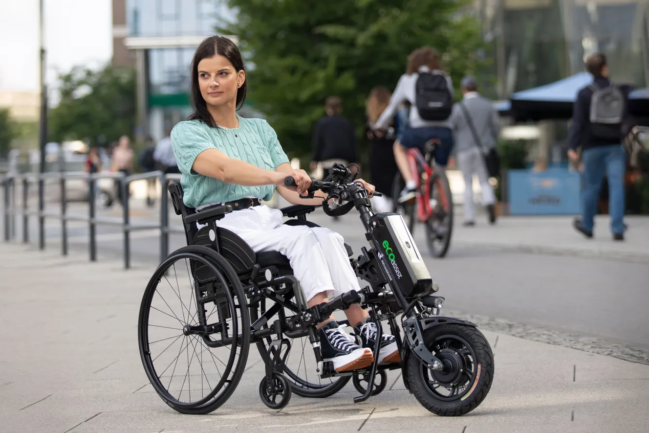 Frau sitzt in Rollstuhl mit eco-assist-Zusatzteil, elektrisch, Vorderrad und Lenker, Straße im Hintergrund