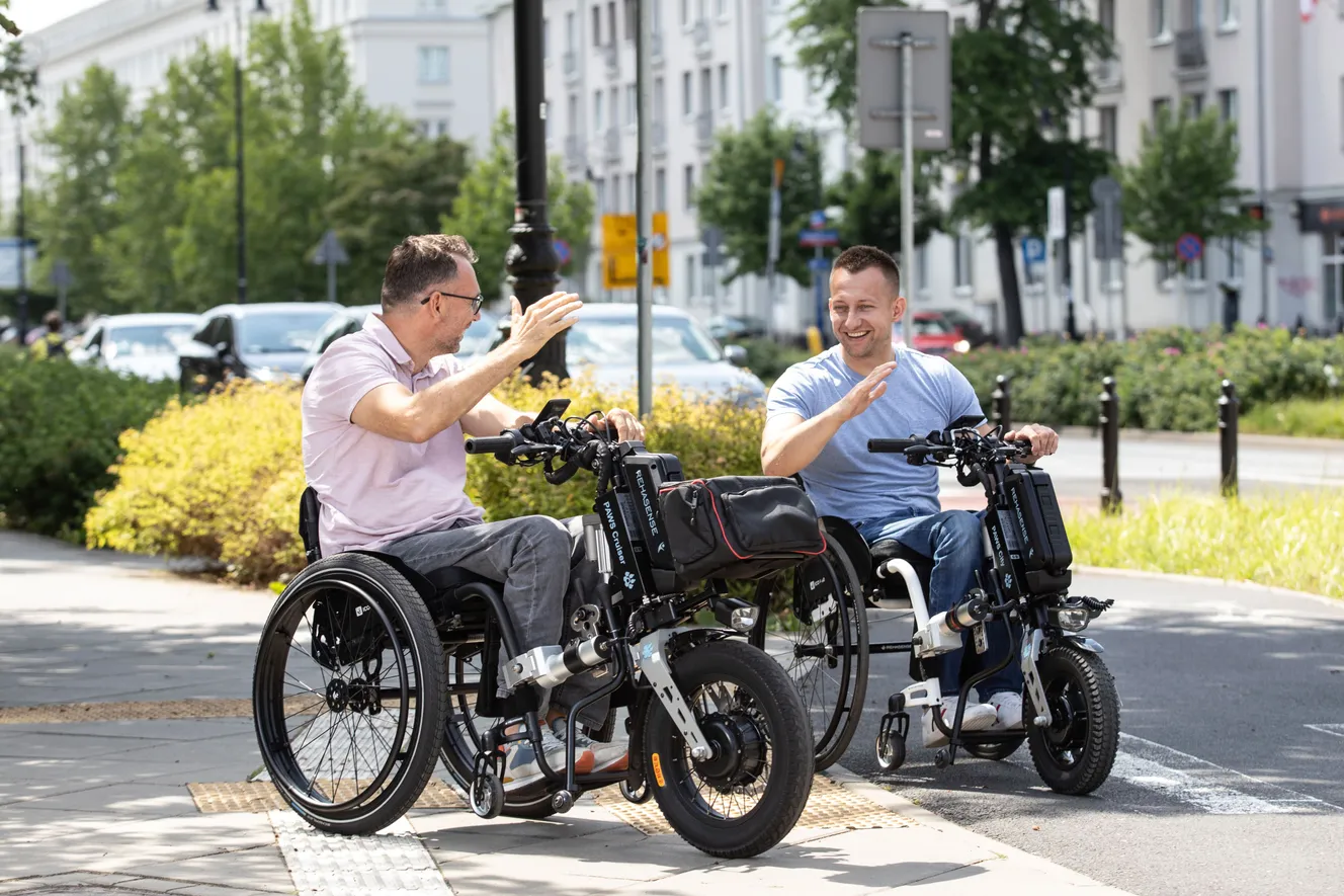 Zwei junge Männer sitzen auf Paws, elektrische Zusatzgeräte für Rollstühle, Vorderrad mit Lenker, schwarz, Straße im Hintergrund