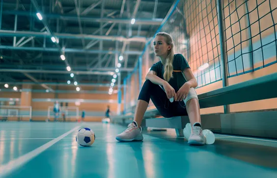 Bänderriss Fuß, sitzende Frau in Sporthalle, ein Fuß wird gekühlt, vor der Frau liegt ein Ball, Sportunfall