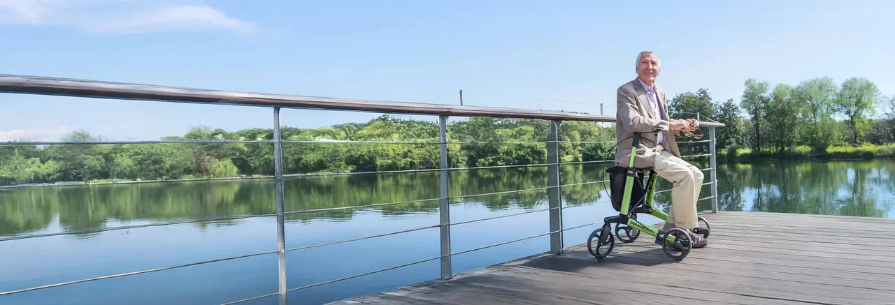 Älterer Mann sitzt auf einem grünen Rollator auf einem Holzsteg über einem ruhigen See