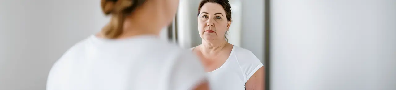 Frau mit Gewichtszunahme bei Lipödem, schaut in den Spiegel, weißes Tshirt