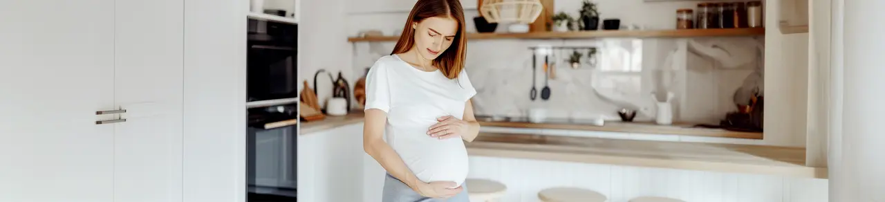 Schwangere Frau mit Symphysenschmerzen hält sich den Bauch, steht in Küche