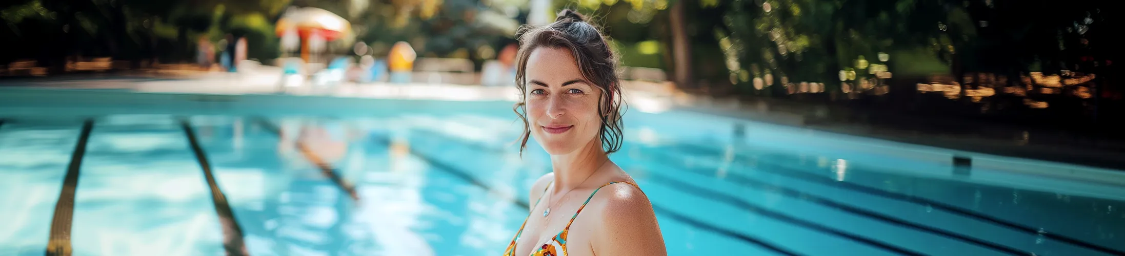 junge Frau mit Brustkrebs steht im Freibad, trägt Badeanzug, lächelt, braune Haare, Pool im Hintergrund