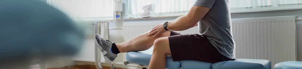 Meniskusriss-Behandlung, Mann sitzt auf Liege, eine Hand am Knie, Physiotherapie