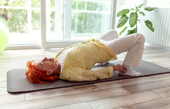 ältere Frau mit Überlaufinkontinenz macht Beckenbodenübung, liegt auf Gymnastikmatte, Fenster, Pflanze, Gymnastikball im Hintergrund