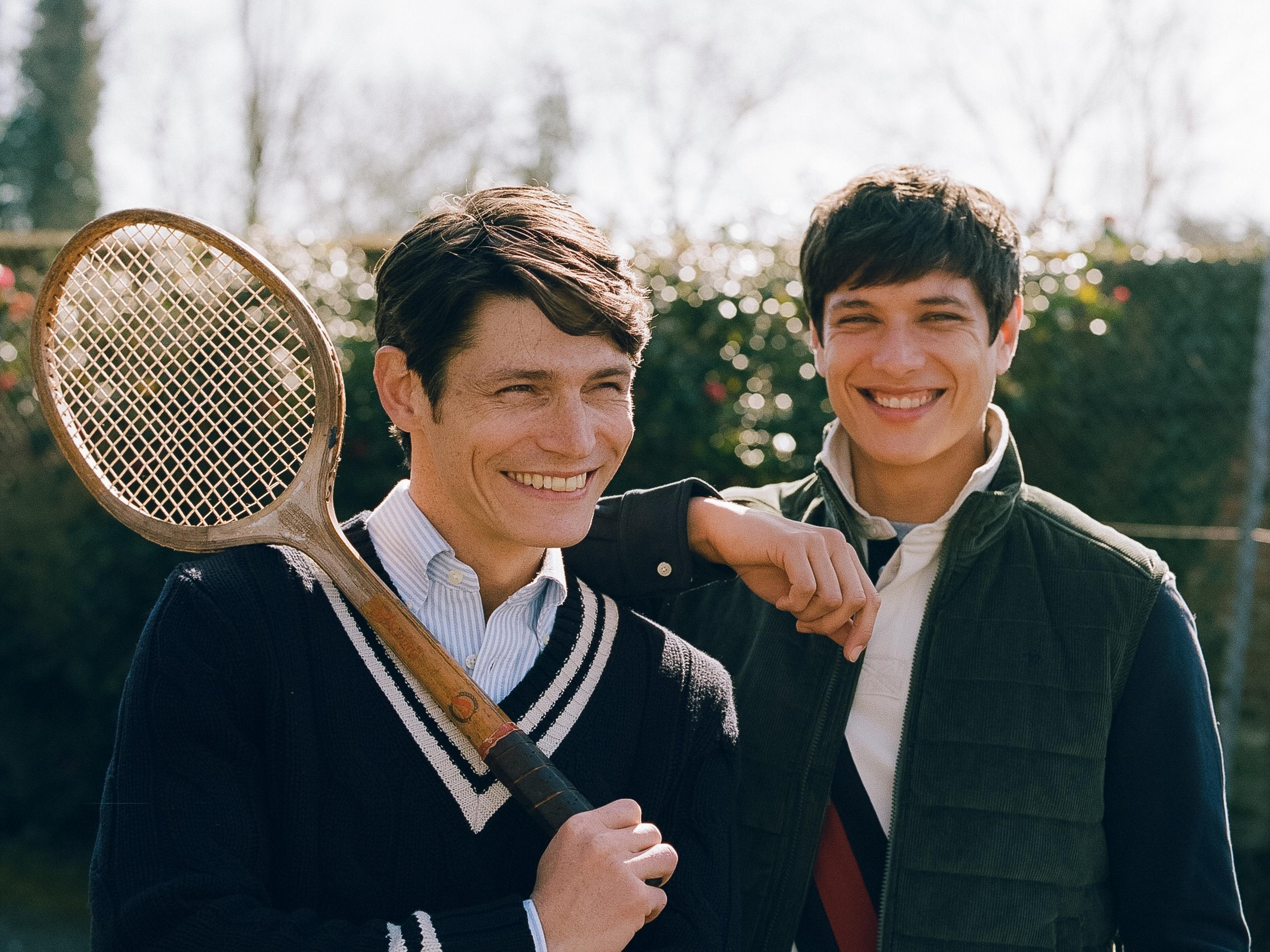 Two men smiling outdoors, one holding a vintage tennis racket. They are dressed in casual, sporty attire with a blurred background of trees and bushes.