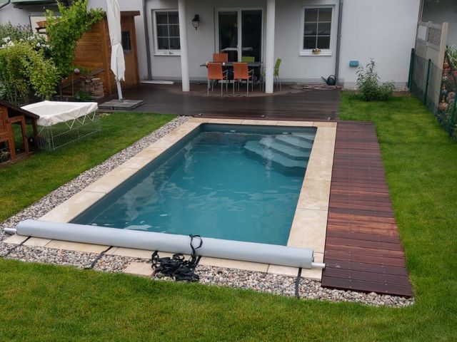Kleiner rechteckiger Swimmingpool in einem Garten hinter einem zweistöckigen Haus mit Balkonen und einer Terrasse, umgeben von grünem Rasen und Kieselsteinen. Auf der Terrasse stehen ein Tisch und Stühle unter einem Sonnenschirm.