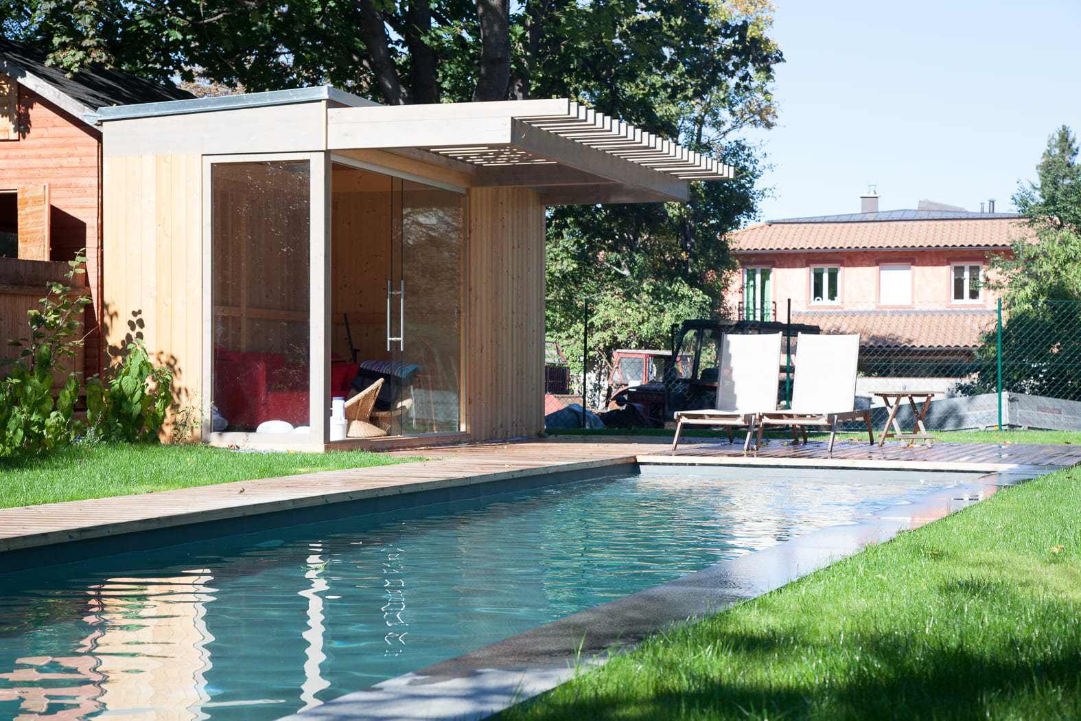 Ein rechteckiger Swimmingpool in einem Garten mit einem modernen Holzpavillon mit Glaswänden, der als Sitzbereich genutzt wird. Neben dem Pool stehen zwei Sonnenliegen auf einem Holzdeck, umgeben von grüner Rasenfläche und Bäumen, mit einem Wohnhaus im Hintergrund.