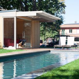 Moderner Gartenpool mit Holzdeck und einem kleinen Gartenhaus im Hintergrund, umgeben von grüner Wiese und Bäumen. Neben dem Pool stehen zwei Sonnenliegen, die eine entspannte Atmosphäre schaffen.