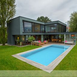 Ein modernes, zweistöckiges Haus mit großen Fenstern und einem flachen Dach. Vor dem Haus befindet sich ein rechteckiger Pool, umgeben von einer Holzterrasse und einer gepflegten Rasenfläche.