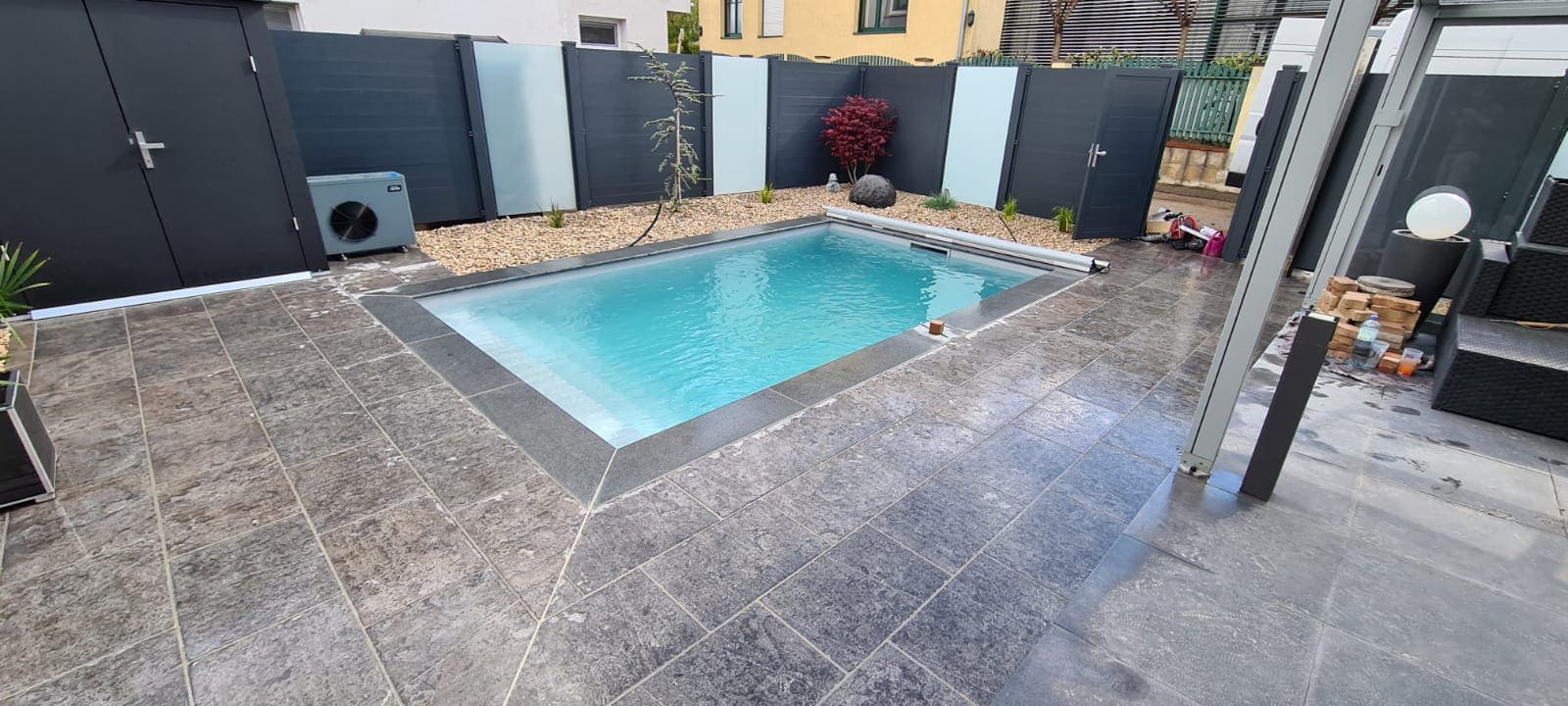 Pool mit grauem Fliesenrand, umgeben von Steinen und modernen Zaunelementen
