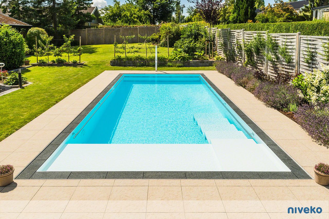 Moderner rechteckiger Pool mit klarem blauem Wasser, umgeben von einer großen Terrasse und gepflegtem Garten mit grünem Rasen und verschiedenen Pflanzen.