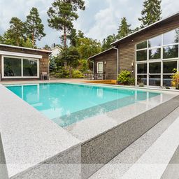 Ein modernes Haus mit großen Fenstern und Holzverkleidung, das sich neben einem eleganten Swimmingpool mit Stufen aus Granit befindet. Der Pool liegt in einem gepflegten Garten, umgeben von Bäumen und üppigem Grün.