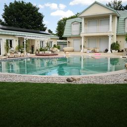 Große, luxuriöse Villa mit grünem Dach und einem großen, geschwungenen Swimmingpool im Vordergrund, umgeben von gepflegtem Rasen und dekorativen Kieselsteinen.