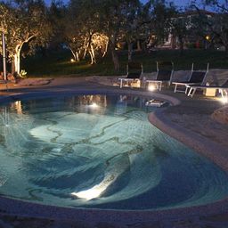 Beleuchteter BiodeEin Biodesign Swimmingpool bei Nacht, beleuchtet von Unterwasserlichtern, umgeben von Liegestühlen und Bäumen. Der Pool befindet sich in einer natürlichen Gartenlandschaft mit sanfter Beleuchtung, die eine entspannte und einladende Atmosphäre schafft.sign Pool erstrahlt am Abend im vollen Glanz.