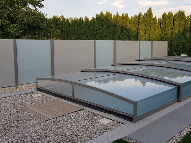 Ein rechteckiger, überdachter Swimmingpool mit einem flachen, verschiebbaren Dach aus Glas und Aluminiumrahmen, umgeben von einem Kiesbett und modernen Sichtschutzwänden. Im Hintergrund sind hohe grüne Hecken und Bäume zu sehen, die die Privatsphäre des Bereichs betonen.