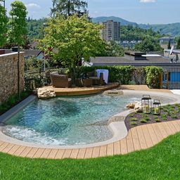 Ein geschwungener, natürlicher Pool mit Holzdeck, grüner Bepflanzung und Felsen, eingebettet in einen gepflegten Garten. Im Hintergrund erstrecken sich städtische Gebäude und Hügel unter einem klaren blauen Himmel.