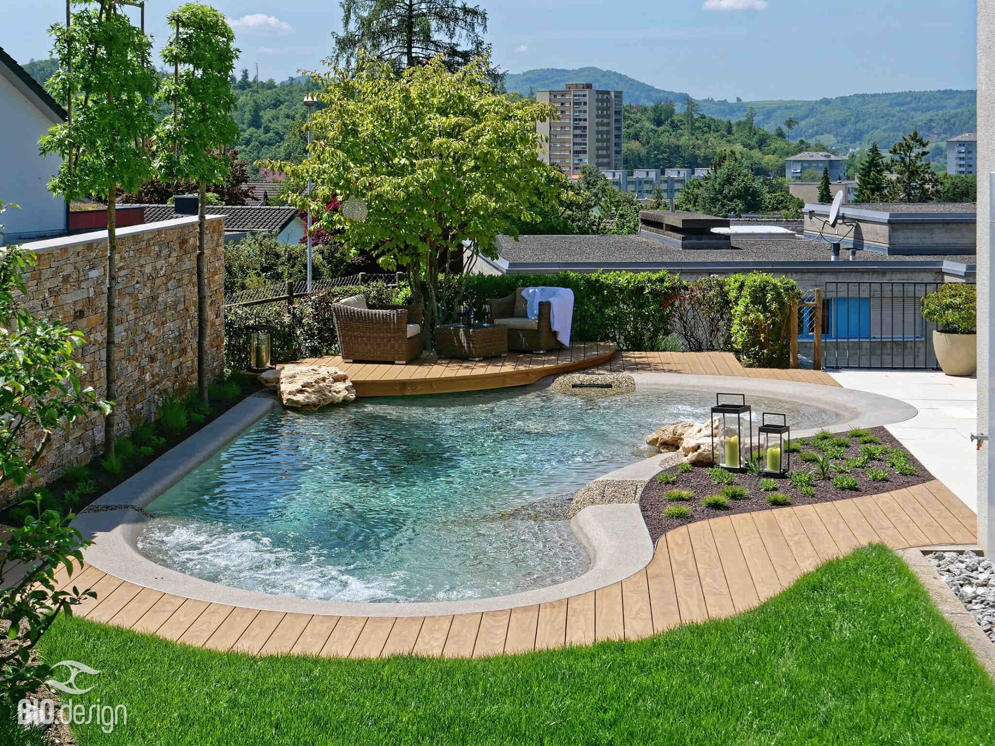Ein eleganter Garten mit einem natürlichen Pool, Holzterrasse, bequemen Sitzmöbeln und Blick auf grüne Hügel und Gebäude.