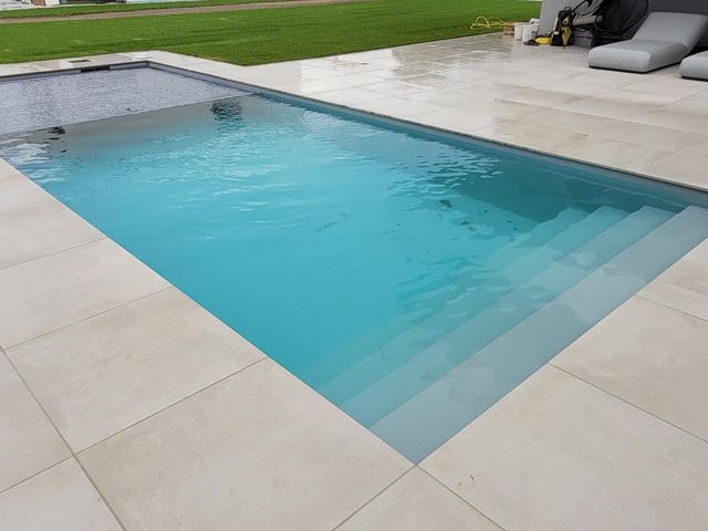 Ein rechteckiger Swimmingpool mit klaren, blauen Wasser, umgeben von einer hellen, gefliesten Terrasse und angrenzender grüner Rasenfläche. Neben dem Pool stehen zwei graue Sonnenliegen.