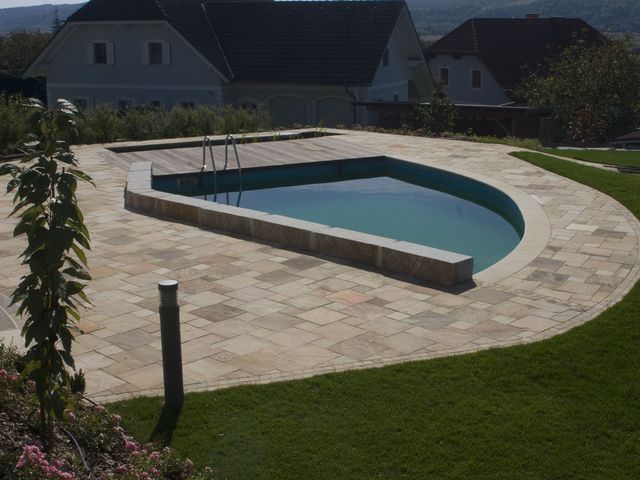 Ein moderner Swimmingpool mit abgerundeten Ecken und integrierter Treppe, umgeben von einem großzügigen, gepflasterten Bereich und gepflegtem Rasen. Im Hintergrund befinden sich Wohnhäuser und eine hügelige Landschaft, die dem Bild eine malerische Kulisse verleihen.