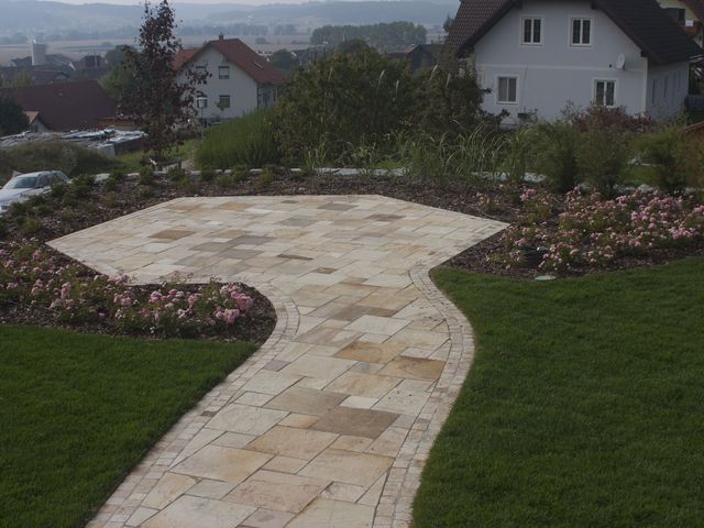 Steinweg in einem gepflegten Garten, der durch blühende Rosenbüsche und grüne Rasenflächen führt. Der Weg biegt sich zu einer gepflasterten Terrasse, die von weiteren Pflanzen und Sträuchern umgeben ist. Im Hintergrund sind mehrere Häuser mit roten und dunklen Dächern zu sehen, sowie ein Auto, das am Rand des Gartens geparkt ist. Die Landschaft im Hintergrund ist hügelig und bietet eine malerische Aussicht.