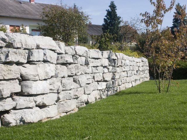 Eine robuste Steinmauer aus großen, unregelmäßig geformten Kalksteinblöcken, die einen gepflegten Garten mit grünem Rasen abgrenzt. Hinter der Mauer ist ein Haus mit einem Schrägdach und einem Garten mit verschiedenen Bäumen und Büschen zu sehen. Ein einzelner Baum mit herbstlichem Laub steht im Vordergrund, während im Hintergrund weitere Pflanzen und Gebäude die ländliche Landschaft prägen.