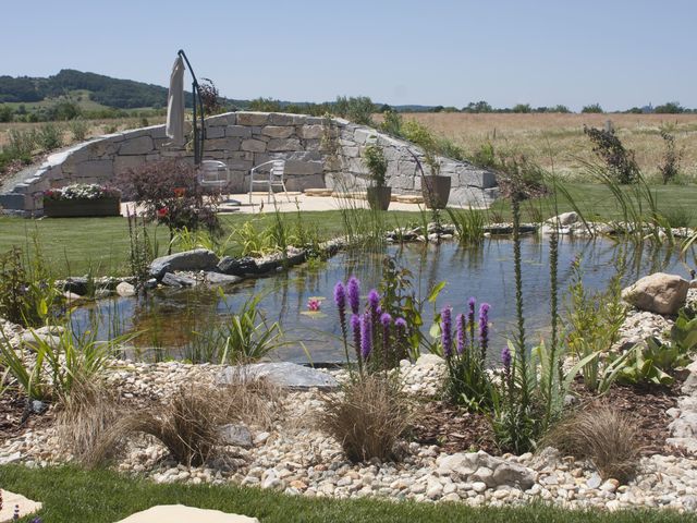Ein Naturteich in einem gepflegten Garten mit blühenden lila Blumen im Vordergrund und einem Sitzbereich aus Stein im Hintergrund. Der Teich ist von verschiedenen Pflanzen umgeben und die Landschaft im Hintergrund besteht aus weiten Feldern und Hügeln.
