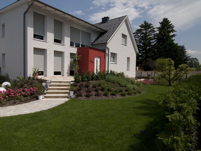Modernes, zweistöckiges Haus mit weißer Fassade und einem roten Eingangsbereich, umgeben von einem gepflegten Garten mit blühenden Blumenbeeten, grüner Rasenfläche und dekorativen Kugelleuchten. Eine Treppe führt vom Hauseingang zur Gartenfläche hinunter, die mit verschiedenen Sträuchern und Pflanzen gestaltet ist.